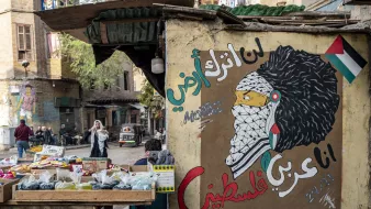 L'image montre un mur peint avec une fresque représentant une figure symbolique, probablement inspirée par la culture palestinienne. Le personnage porte un keffieh et a une chevelure élaborée. À côté, il y a des inscriptions en arabe qui expriment des sentiments de résistance et d'identité palestinienne. En avant-plan, il y a un marché avec des étals de fruits et légumes, et une rue animée où l'on peut apercevoir des passants et des véhicules. L'ambiance reflète un mélange de culture, d'art et de vie urbaine.