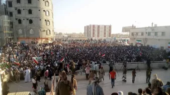 L'image montre une grande foule rassemblée lors d'une manifestation ou d'un rassemblement public. Les participants tiennent des banderoles et des drapeaux, indiquant un fort engagement politique ou social. En arrière-plan, on peut voir des bâtiments en construction ou rénovés, ainsi qu'une architecture variée. La scène semble se dérouler en plein air, probablement durant l'après-midi ou le début de la soirée, avec une lumière naturelle éclatante. L'atmosphère est dynamique, avec beaucoup de gens engagés dans une cause commune.