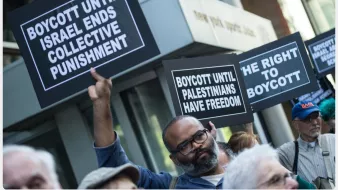 L'image montre une manifestation avec des personnes tenant des pancartes. Les pancartes portent des messages appelant au boycott jusqu'à ce qu'Israël mette fin à des mesures de punition collective et jusqu'à ce que les Palestiniens aient la liberté. On peut voir des participants de différents âges, tous unis par des revendications similaires. L'arrière-plan indique un environnement urbain avec des bâtiments.