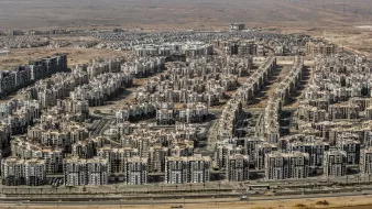 L'image montre une vaste zone urbaine composée de nombreux immeubles de plusieurs étages, organisés de manière dense. On peut voir des routes qui traversent ces secteurs résidentiels, avec des infrastructures modernes. En arrière-plan, le paysage est désertique, ce qui évoque un contraste entre l'urbanisation et l'environnement naturel environnant. L'ensemble dégage une impression de développement urbain important dans une région aride.