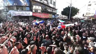 L'image montre une grande foule rassemblée dans une rue, avec des personnes portant des drapeaux et des pancartes. Il y a des cercueils sur des supports, symbolisant un hommage ou des funérailles. La foule est visiblement très engagée, levant les mains et exprimant des émotions fortes. L'environnement urbain est marqué par des bâtiments et des affiches en arrière-plan. L'atmosphère est chargée de gravité et de solidarité.
