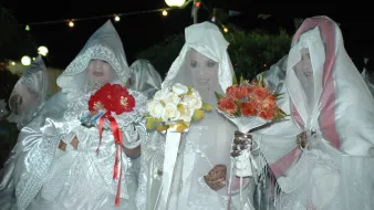 L'image montre un groupe de femmes vêtues de robes blanches et de voiles, se tenant ensemble dans une ambiance nocturne. Elles tiennent chacune des bouquets de fleurs, composés de roses et d'autres fleurs colorées. L'éclairage doux crée une atmosphère festive, et des guirlandes lumineuses sont visibles en arrière-plan. Leurs visages sont en partie cachés par les voiles, ajoutant une aura de mystère à la scène.