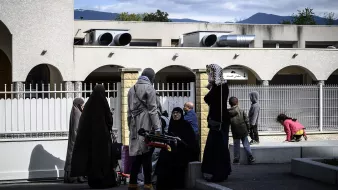 L'image montre un groupe de personnes à l'extérieur d'un bâtiment. Certaines femmes portent des vêtements longs et des foulards, tandis que d'autres semblent habillées plus décontractées. On aperçoit également des enfants jouant dans l'arrière-plan. Le ciel est partiellement nuageux et des montagnes se dessinent au loin. L'ambiance semble conviviale, avec des échanges entre les personnes présentes.