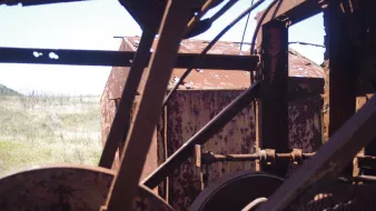 L'image montre une vue d'une structure métallique, probablement une machine ou un équipement abandonné, avec des surfaces rouillées et en décomposition. On peut voir des éléments mécaniques, tels que des poulies ou des engrenages, qui sont partiellement visibles à travers des parties de la machine. En arrière-plan, on distingue un paysage de prairies sèches, ce qui évoque une ambiance d'abandon et de dégradation. La lumière naturelle met en valeur les textures rouillées du métal.