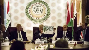 L'image montre une réunion officielle, probablement dans le cadre d'une organisation internationale. Plusieurs hommes sont assis autour d'une table dans un espace bien décoré, avec des drapeaux de différents pays en arrière-plan. L'un des hommes, au centre, tient un document ou un graphique qu'il montre aux autres participants. Les autres semblent attentifs et engagés dans la discussion. Les éléments visuels suggèrent une atmosphère sérieuse et formelle, en lien avec des discussions diplomatiques.