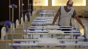 L'image montre une personne qui se trouve dans un environnement médical, probablement un hôpital ou un centre de soins. Cette personne porte un masque et un gilet, et elle est en train de s'occuper de plusieurs lits médicaux alignés. Les lits sont équipés de draps et de dispositifs intraveineux. L'atmosphère semble calme et organisée, suggérant un espace dédié aux soins des patients.
