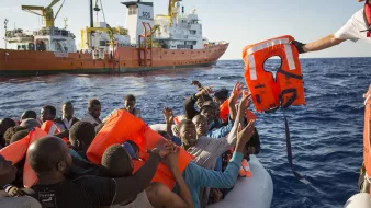 L'image montre une scène en mer où un groupe de personnes se trouve sur un canot pneumatique. Elles semblent en détresse et soulèvent les bras en direction d'un sauveteur qui leur tend une bouée de sauvetage. En arrière-plan, un grand navire de secours est visible, avec des couleurs orange et blanc. L'atmosphère semble tendue, et on peut ressentir l'urgence de la situation. Le ciel est dégagé et l'eau est calme, contrastant avec la gravité du moment.