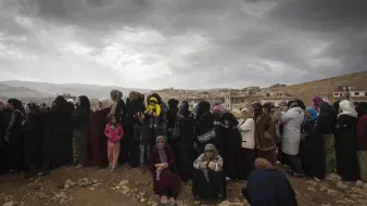 L'image montre un groupe de femmes rassemblées, probablement en attente de quelque chose. Elles portent des vêtements sombres, et certaines tiennent des enfants. Le ciel est nuageux, ce qui donne une ambiance sombre à la scène. En arrière-plan, on aperçoit des maisons sur une colline, soulignant un environnement rural ou de village. L'attente et la solidarité féminine semblent être des thèmes présents dans cette image.