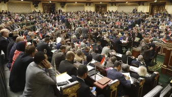 L'image montre une assemblée ou un parlement en pleine session. De nombreux représentants sont assis dans des rangées, certains discutent tandis que d'autres prennent des notes ou consultent des documents. On peut voir des drapeaux égyptiens sur certaines tables, ce qui suggère que cette image provient de l'Assemblée nationale d'Égypte. L'environnement est formel, avec une riche décoration et une disposition typique d'une salle de réunion politique.