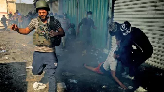 L'image montre une scène chaotique et tendue, probablement lors d'une manifestation ou d'un événement violent. Au premier plan, un homme portant un casque de protection et un gilet semble fuir rapidement, tandis qu'en arrière-plan, d'autres personnes sont engagées dans une altercation. L'atmosphère est remplie de fumée, ce qui ajoute à la tension de la situation. Le sol est jonché de débris, suggérant que l'incident a eu lieu dans un environnement urbain. Des gens semblent être en détresse, reflétant l'intensité du moment.