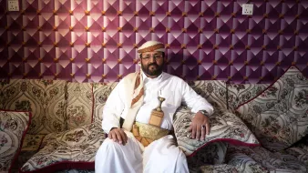 L'image montre un homme vêtu d'un habit traditionnel, assis sur un canapé orné de motifs décoratifs. Il porte une ceinture dans laquelle est insérée une sorte de poignard, typique de certaines cultures arabes. Le fond est constitué d'un mur avec un revêtement texturé en violet, ajoutant une touche colorée à la scène. L'homme a une expression calme et posée, et son environnement reflète une atmosphère accueillante et traditionnelle.