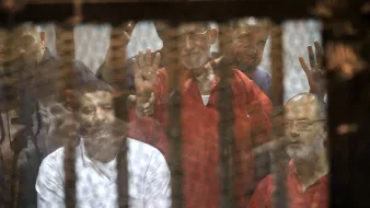 L'image montre un groupe d'hommes derrière une grille ou un panneau de verre. Ils semblent être dans un contexte de détention ou de procès, avec des expressions variées sur leurs visages. Certains lèvent les mains, suggérant un geste de protestation ou de solidarité. L'ambiance générale est sombre, évoquant des thèmes de justice et de droits humains.