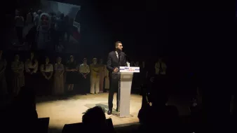 L'image montre un homme debout à un pupitre transparent, prenant la parole lors d'un événement. Il est habillé en costume et semble s'adresser à un public. En arrière-plan, on peut voir un écran projetant une vidéo ou une image, vraisemblablement liée à un sujet important, étant donné l'expression des personnes présentes. Au premier plan, on peut apercevoir des membres du public et des photographes, créant une atmosphère de concentration et d'engagement. L'éclairage met en évidence l'orateur, tandis que le reste de la scène est plus sombre.