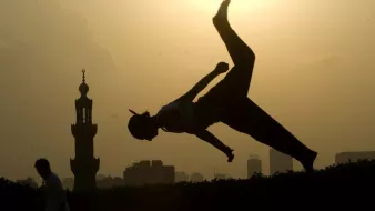 L'image montre une silhouette d'une personne effectuant un saut ou un mouvement acrobatique contre un fond de soleil couchant. À l'arrière-plan, on aperçoit une mosquée avec un minaret, ainsi que des bâtiments de la ville. L'éclairage crée un contraste fort entre la silhouette et le ciel lumineux, ajoutant une atmosphère dynamique et artistique à la scène.