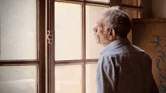 L'image montre un homme âgé qui semble contempler à travers une fenêtre. Il est habillé d'une chemise à motifs et a une posture pensive. La lumière naturalisée entre par la fenêtre, illuminant doucement son visage et créant une ambiance calme. On peut aussi apercevoir des murs en arrière-plan, ajoutant à l'atmosphère de réflexion du moment.