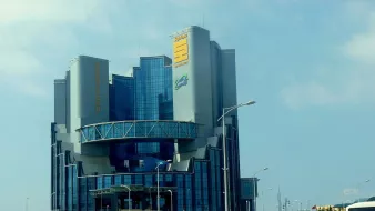 L'image montre un bâtiment moderne et imposant, avec une architecture contemporaine. La structure se dresse sur plusieurs niveaux, avec de grandes fenêtres en verre qui reflètent la lumière. Au sommet, il y a un panneau avec des inscriptions en caractères distincts, suggérant que le bâtiment abrite une entreprise ou une organisation importante. Il est entouré d'une route et d'un espace vert, ce qui ajoute au cadre urbain. Le ciel est partiellement nuageux, ce qui donne un éclairage intéressant à l'ensemble de la scène.