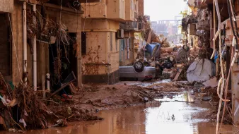 L'image montre une rue dévastée par une inondation. On peut voir des bâtiments endommagés, avec des débris et de la boue accumulés sur le sol. Des voitures sont renversées et se trouvent parmi les débris, tandis qu'une faible couche d'eau stagnante reflète la destruction environnante. L'atmosphère est sombre, soulignant les conséquences dramatiques de la catastrophe.