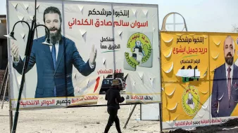 L'image montre un homme marchant devant de grandes affiches électorales dans un environnement urbain. Les affiches présentent des candidats politiques avec leurs noms écrits en arabe, ainsi que des logos et des slogans associés à leur campagne. L'arrière-plan semble désordonné, avec des structures inachevées ou des éléments typiques d'une ville. L'homme semble porter une charge sur sa tête, ajoutant une dimension de vie quotidienne à la scène.