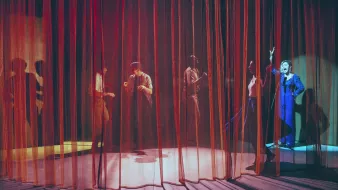 L'image montre une scène de théâtre avec des rideaux en velours rouge et orange qui créent une atmosphère intimiste. On aperçoit plusieurs silhouettes de personnes derrière les rideaux, engagées dans des activités qui semblent dynamiques et expressives. L'un des personnages, visible à droite, porte un costume bleu et semble gesticuler avec enthousiasme. Les jeux de lumière entre les rideaux ajoutent une ambiance dramatique à la scène, accentuant les mouvements des acteurs.