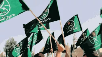 L'image montre un groupe de personnes levant des drapeaux verts. Les drapeaux semblent porter des inscriptions en arabe ainsi que des symboles, indiquant une affiliation à un mouvement ou une organisation spécifique. L'ambiance semble dynamique et engagée, suggérant un rassemblement ou une manifestation. L'arrière-plan indique probablement un espace public ou un lieu symbolique.
