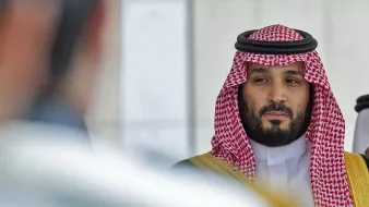 L'image montre un homme portant des vêtements traditionnels saoudiens, y compris un keffieh et un thobe. Il a une expression sérieuse et regarde vers la caméra ou un point de vue adjacent. Son entourage semble formel, avec une ambiance de cérémonie ou de rencontre officielle. Les couleurs dominantes sont les tons de rouge et de blanc du keffieh, ainsi que des touches dorées sur sa tenue.