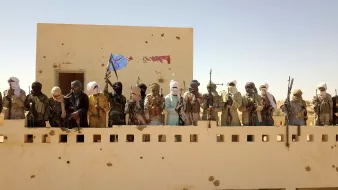 L'image montre un groupe de personnes armées, probablement des membres d'un groupe militant, se tenant sur un balcon ou une terrasse d'un bâtiment. Ils portent des vêtements traditionnels et des masques, et certains brandissent des armes. Le fond est désertique et ensoleillé, avec un ciel clair. On remarque également un drapeau dans le groupe, suggérant un certain degré d'organisation ou d'identité collective.