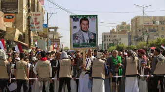 L'image montre une manifestation dans une rue animée, avec une foule de personnes rassemblées. Au premier plan, il y a un groupe de manifestants portant des vêtements traditionnels et des gilets beige. Ils se tiennent derrière une barrière et semblent faire face à l'objectif. En arrière-plan, on voit des drapeaux, ainsi qu'une grande affiche représentant une personne, probablement un leader ou une figure importante. L'ambiance semble être chargée d'émotion et de détermination, avec un secteur urbain typique visible autour.