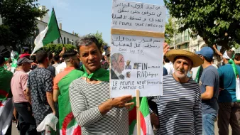 L'image montre un groupe de personnes manifestant, principalement des hommes, certains portant des drapeaux algériens. Deux manifestants se tiennent en avant-plan, l'un tenant une pancarte avec des inscriptions en arabe et en français. La pancarte semble exprimer des revendications politiques, notamment la fin de certaines pratiques liées à la politique algérienne. Les manifestants semblent unis et déterminés, se rassemblant pour faire entendre leur voix. L'atmosphère générale est celle d'une protestation pacifique.