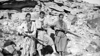 L'image montre trois hommes vêtus de vêtements traditionnels, tenant chacun un fusil. Ils se tiennent debout sur un sol rocheux, entourés de grandes pierres et de formations rocheuses. L'environnement semble austère et désertique. Les hommes ont des expressions sérieuses et sont alignés côte à côte. Leur posture et l'équipement qu'ils portent suggèrent qu'ils peuvent être des membres d'un groupe armé ou des guerriers appartenant à une communauté spécifique.
