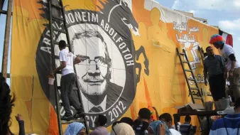 L'image montre un groupe de personnes en train de peindre une grande bannière ou une affiche. Ils sont sur des échelles et travaillent à mettre en place un portrait ou un dessin qui semble représenter une figure importante, peut-être liée à un événement politique. Le fond est coloré, principalement en orange, et il y a des inscriptions en arabe. Les femmes au premier plan portent des foulards, et l'atmosphère semble dynamique, avec des gens engagés dans le travail collectif.