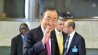 L'image montre un homme souriant, vêtu d'un costume sombre et d'une cravate rose, qui fait un geste de la main. Il semble être dans un environnement formel, éventuellement un bâtiment ou une salle de conférences. En arrière-plan, on distingue d'autres personnes en costume, donnant une impression d'un événement ou d'une réunion officielle. L'homme au premier plan pourrait être une figure publique, mais je ne peux pas en dire plus sur son identité.