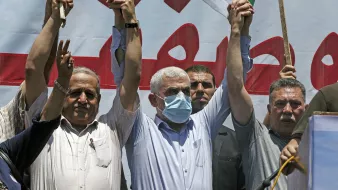 L'image montre un groupe de personnes levant les bras en signe de solidarité ou de protestation. Certains portent des masques, et il y a des drapeaux visibles en arrière-plan. L'ambiance semble être celle d'un rassemblement ou d'une manifestation. Les expressions des participants varient, reflétant des émotions de détermination ou de franche camaraderie.