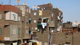 L'image montre un paysage urbain d'un quartier populaire. On peut voir des bâtiments en briques de terre, souvent de plusieurs étages, avec des façades colorées. De nombreuses antennes parabolique sont installées sur les toits. Les vêtements sont accrochés à l'extérieur pour sécher. L'atmosphère semble animée, avec des signes de vie quotidienne. Le ciel est dégagé, ce qui donne une impression de chaleur.