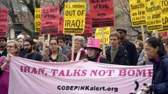 L'image montre une manifestation où des personnes tiennent des pancartes avec des slogans anti-guerre. Plusieurs affiches demandent le retrait des troupes des États-Unis d'Irak, de Syrie et d'Afghanistan. Les manifestants semblent déterminés et unis dans leur message, qui appelle à la paix, avec un grand banner rose affichant "IRAN, TALKS NOT BOMBS" et le site web de CODEPINK. L'atmosphère semble à la fois sérieuse et engagée.