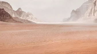 L'image montre un vaste paysage désertique. On peut voir des montagnes rocheuses au loin, entourées de dunes de sable. Le ciel est nuageux, ce qui crée une atmosphère légèrement brumeuse. Au centre, une seule silhouette humaine, probablement un cavalier, se déplace à travers ce paysage aride. La scène transmet une sensation d'immensité et de solitude.