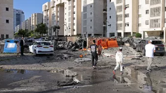L'image montre une scène de destruction urbaine. On peut voir plusieurs véhicules endommagés, certains complètement détruits. Le sol est couvert de débris, et il semble qu'une explosion ou un événement violent ait eu lieu. En arrière-plan, il y a des bâtiments modernes. Des personnes marchent sur la route, apparemment en train d'évaluer les dégâts. Le ciel est clair, ce qui contraste avec l'image de désolation.