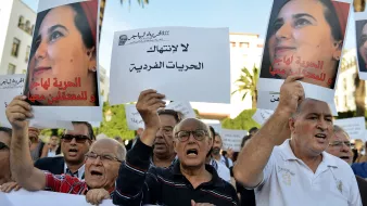 L'image montre une manifestation avec un groupe de personnes tenant des pancartes. Les manifestants semblent exprimer leur désaccord et revendiquer des droits, notamment en ce qui concerne les libertés individuelles. Certains affichent des expressions de colère ou de détermination, tandis que d'autres portent des signes clairs de solidarité. En arrière-plan, on peut voir des palmiers et un bâtiment, suggérant un cadre urbain. L'atmosphère est engagée et militante, reflétant une lutte pour des causes sociales ou politiques.
