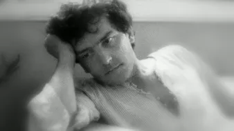 L'image montre un homme pensif, assis dans un endroit confortable. Il a les cheveux bouclés et un air réfléchissant, avec une main soutenant sa tête. Son expression semble mélancolique ou pensive. L'éclairage doux donne une ambiance nostalgique et calme à la scène. L'arrière-plan flou contribue à centrer l'attention sur son visage.