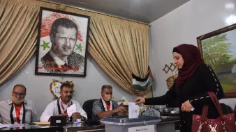 L'image montre une scène de vote, probablement lors d'élections en Syrie. Au premier plan, une femme en hijab dépose un bulletin dans une urne transparente. À l'arrière-plan, plusieurs hommes sont assis derrière une table, regardant le processus. Un grand portrait d'un homme, probablement un dirigeant, est accroché au mur, entouré de drapeaux et de décorations. Les participants portent des badges d'identification, et l'atmosphère semble sérieuse et formelle.