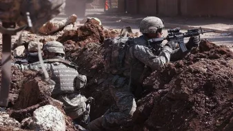 L'image montre des soldats en engagement militaire, se cachant derrière des tranchées. Ils portent des uniformes militaires et des casques, et sont armés. Le terrain est marqué par des gravats et de la terre, suggérant un environnement de combat. L'atmosphère est tendue, montrant la préparation et la vigilance des soldats face à un danger.