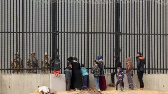 L'image montre un groupe de personnes se tenant devant une grande clôture métallique, probablement une barrière de sécurité. De l'autre côté, des soldats en uniforme semblent observer le groupe. Les personnes sont composées de familles et d'enfants, certains d'eux essaient de communiquer ou de se rapprocher de la clôture. Le cadre est marqué par un environnement désertique, avec des barbelés visibles au sol. L'atmosphère semble chargée d'émotion, soulignant une séparation entre les civils et les forces de sécurité.