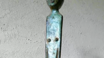 L'image montre un objet en métal, probablement en bronze, qui semble être une sorte de sculpture ou de barre. La partie supérieure représente une tête humaine stylisée, tandis que la tige qui descend présente des éléments qui pourraient évoquer un corps. La surface a une patine verdâtre, indiquant son ancienneté. L'ensemble de l'objet évoque une forme artistique, fusionnant des éléments figuratifs et abstraits.