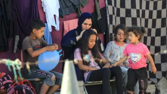 L'image montre un groupe d'enfants et une femme assis à l'extérieur, dans un environnement simple. Les enfants semblent interagir joyeusement, avec une petite fille qui parle à une autre. Une femme en vêtements traditionnels s'occupe des cheveux d'une jeune fille assise devant elle. En arrière-plan, on peut voir des vêtements accrochés et un sol en sable. L'ambiance est conviviale et émotive, évoquant des moments de partage et de complicité.