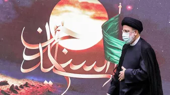 L'image montre un homme en tenue traditionnelle, portant un turban et un masque, qui s'approche d'un fond avec des éléments graphiques. Ce fond présente également une grande lune et un drapeau vert, ainsi que des inscriptions en arabe. L'atmosphère semble solennelle et festive, probablement liée à un événement religieux ou culturel.