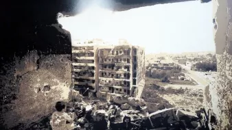 L'image montre l'intérieur d'un bâtiment en ruine, avec un trou dans le mur par lequel on peut voir un paysage extérieur. On aperçoit des bâtiments endommagés au loin, témoignant d'une destruction significative. Le sol est jonché de débris, et l'éclairage semble mettre en évidence la désolation des lieux, créant une atmosphère sombre et mélancolique.