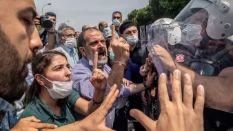 L'image montre une scène de tension entre des manifestants et des forces de l'ordre. Au premier plan, plusieurs personnes expriment leur désaccord, avec des gestes vifs et des expressions de colère ou de frustration. Certaines personnes portent des masques, probablement en raison de préoccupations sanitaires. En arrière-plan, des membres des forces de l'ordre, équipés de casques et de boucliers, semblent faire face aux manifestants. L'environnement est urbain, et le ciel est partiellement nuageux. L'atmosphère est chargée d'émotion et de conflit.