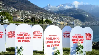 L'image montre un rassemblement commémoratif avec plusieurs pierres tombales blanches disposées sur un terrain vert. Chaque pierre tombale est marquée de rouge avec des inscriptions telles que "More than 100,000 lives lost in Syria". À côté des pierres tombales, il y a des roses ou des plantes, symbolisant probablement la mémoire des vies perdues. En arrière-plan, on peut voir une vue magnifique de montagnes et d'une ville au bord de l'eau. L'ambiance générale semble être solennelle et réfléchissante, soulignant une tragédie humanitaire.