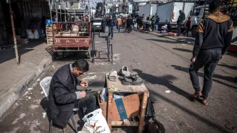 L'image montre une scène de rue animée. Au premier plan, un homme est assis sur une chaise, concentré sur sa tâche. Il répare des chaussures, utilisant des outils comme des ciseaux et des filaments. Devant lui, une table supporte des chaussures à réparer et un sac. À l'arrière-plan, on peut voir des personnes marchant le long d'une rue entourée de camions et de petites échoppes. L'atmosphère semble vivante, avec des éléments de la vie quotidienne qui se déroulent autour de lui.