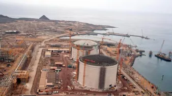 L'image montre un site industriel en construction, probablement une installation de gaz naturel ou une centrale électrique. On peut voir plusieurs grands réservoirs cylindriques, entourés de grues et de matériaux de construction. L'environnement est côtier, avec une vue sur la mer et des montagnes en arrière-plan. Il y a également des routes et des véhicules de construction présents sur le site. Le paysage est principalement désertique et montagneux.