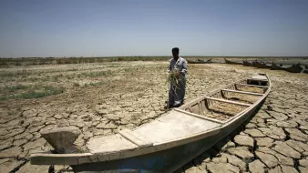 L'image montre un homme debout sur un sol craquelé, typique d'une région aride. Il semble tenir une poignée de végétation, peut-être de l'herbe ou des tiges. À ses pieds se trouve une barque vide, qui est en partie enfouie dans le sol sec. En arrière-plan, on peut apercevoir d'autres barques, mais le paysage aride, sans présence significative d'eau, souligne un environnement désolé et dégradé. Le ciel est dégagé et lumineux, renforçant l'atmosphère de chaleur et de sécheresse.